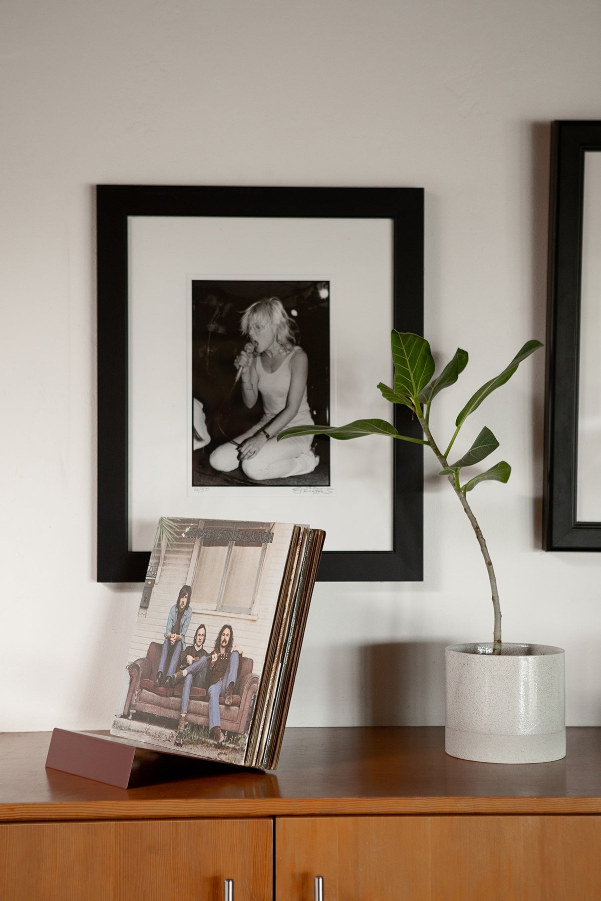 Upton LP Stand in rose (pink/red). Modern and minimalist tabletop steel vinyl record holder made for storage, organization, and display of 50 albums. Shown holding a stack of records with a black and white photo on a wall with a plant and books on a wooden surface below, in a modern interior.