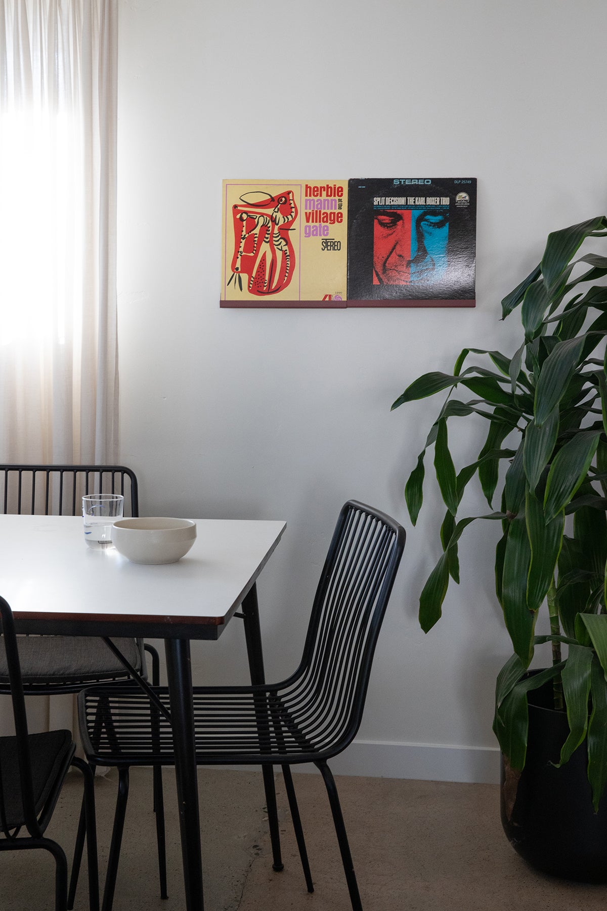 Upton LP Wall Display in black. Modern and minimalist steel wall shelf made to hang vinyl record sleeves on a wall. Shown holding two albums in a dining area with a table, chairs, and plant in a modern interior.
