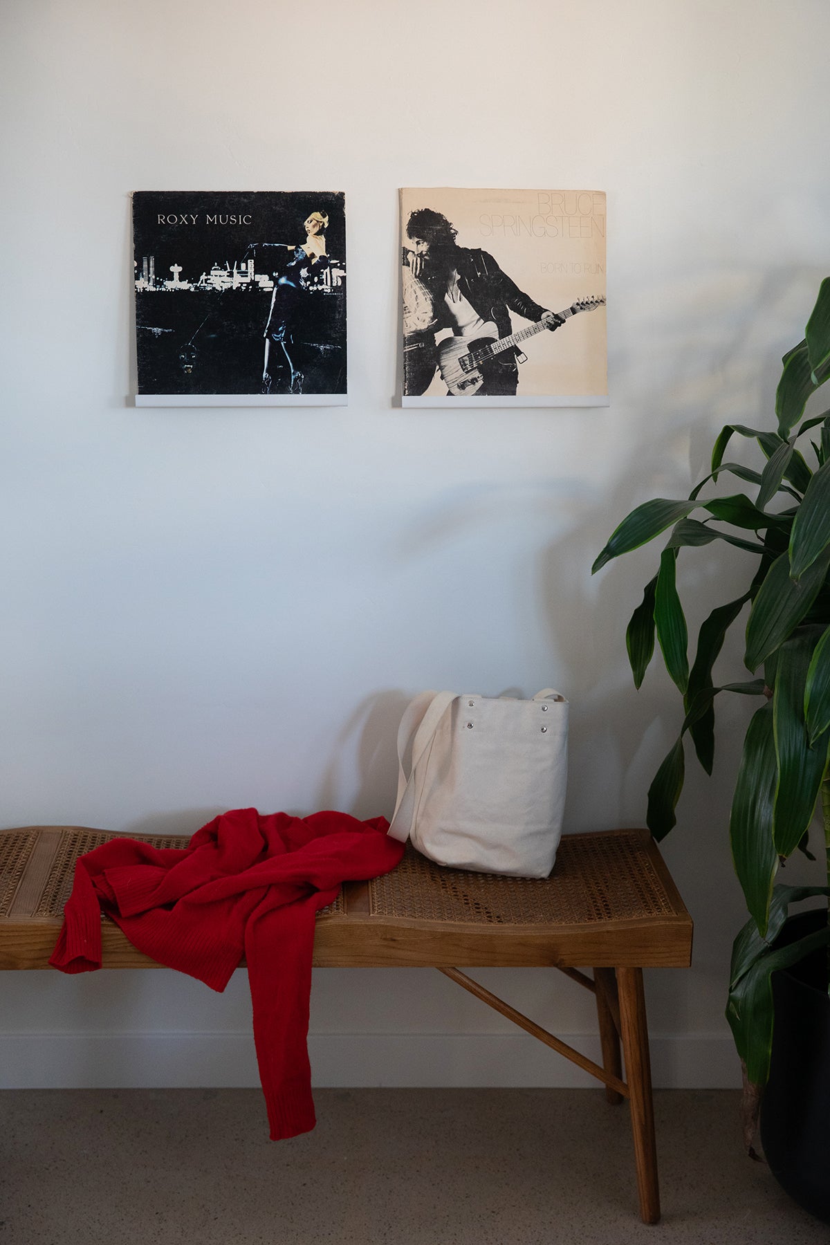 Upton LP Wall Display in white. Modern and minimalist steel wall shelf made to hang vinyl record sleeves on a wall. Shown holding two Roxy Music albums on a white wall over a wooden bench with white bag and red sweater.