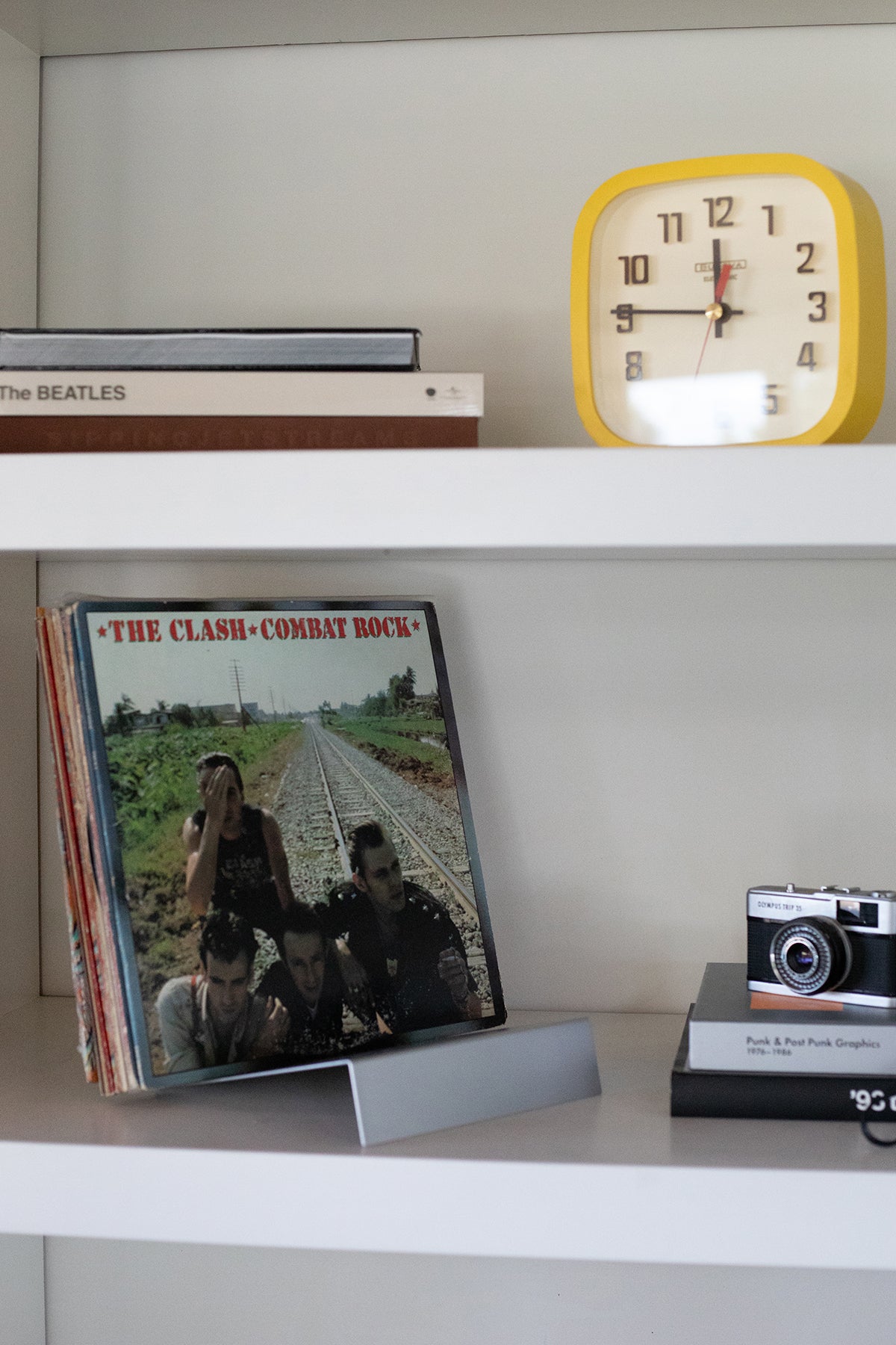Upton LP Stand in matte silver. Modern and minimalist tabletop steel vinyl record holder made for storage, organization, and display of 50 albums. Shown holding a stack of albumbs with 'The Clash - Combat Rock' on top on a shelf with a clock and camera model.