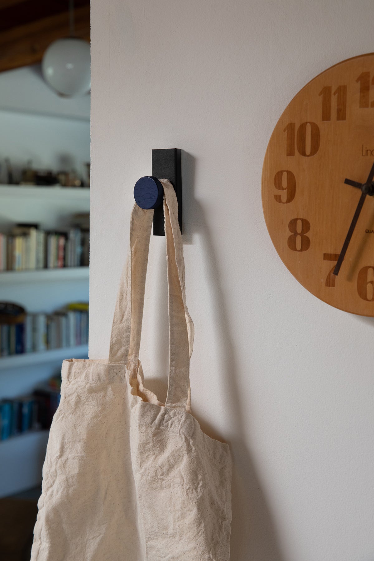 Upton Canal Wall Hook in black/navy. A sculptural decorative steel and wood wall hook with a black and navy blue color scheme with a unique shape, designed to hang bags, coats, towels, hats, and more. Shown here holding a cream tote next to a wooden clock.