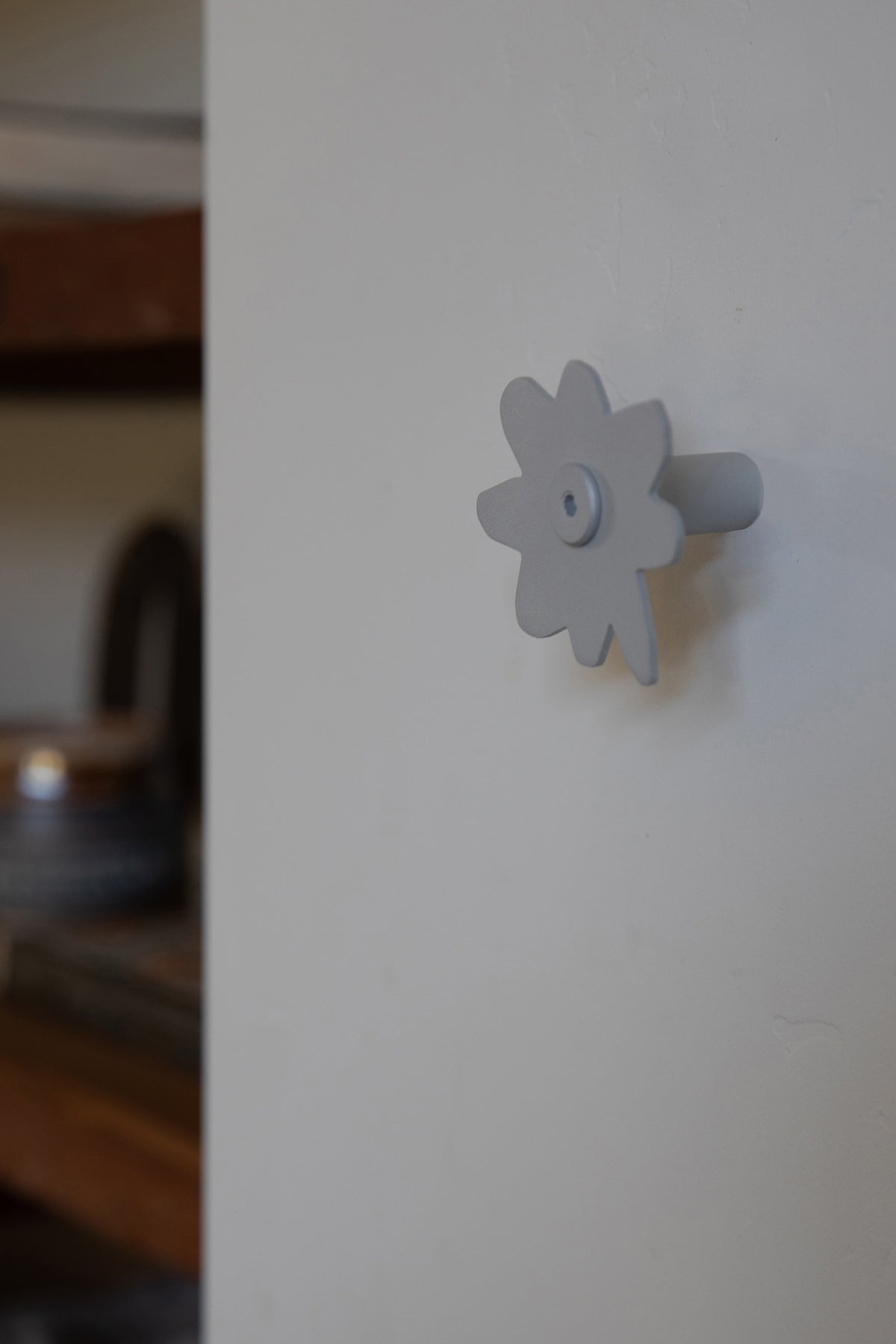 Upton Fleur Wall Hook in matte silver. A sculptural decorative steel wall hook with a silver color scheme with a unique abstract flower shape, designed to hang bags, coats, towels, hats, and more. Shown here mounted on a white wall in a rustic interior with blurred open wooden shelving in the background.