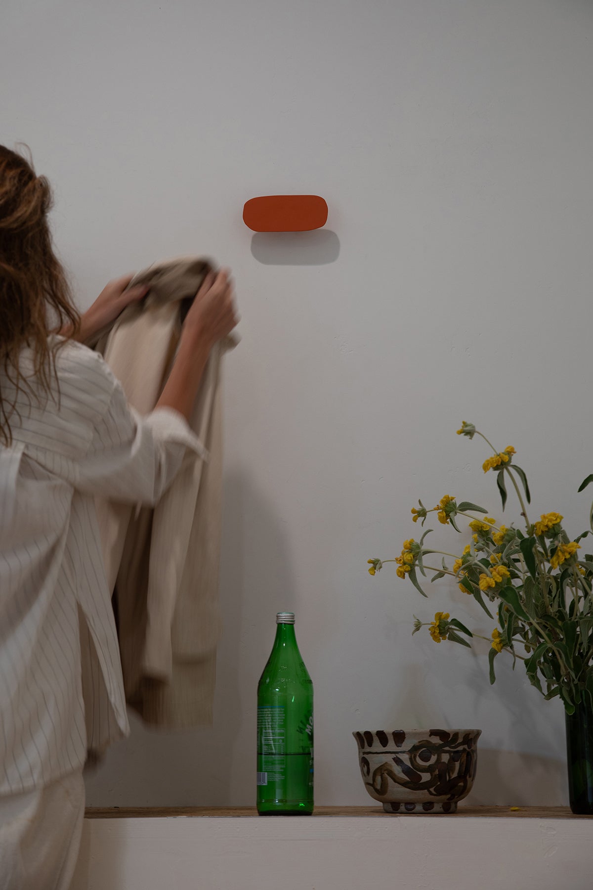 Upton Pill Wall Hook in burnt orange. A sculptural decorative steel wall hook with a burnt orange color scheme with a unique abstract shape, designed to hang bags, coats, towels, hats, and more. Shown here on a white wall with a woman hanging a coat on it, with a green bottle and handmade bowl and vase of flowers.