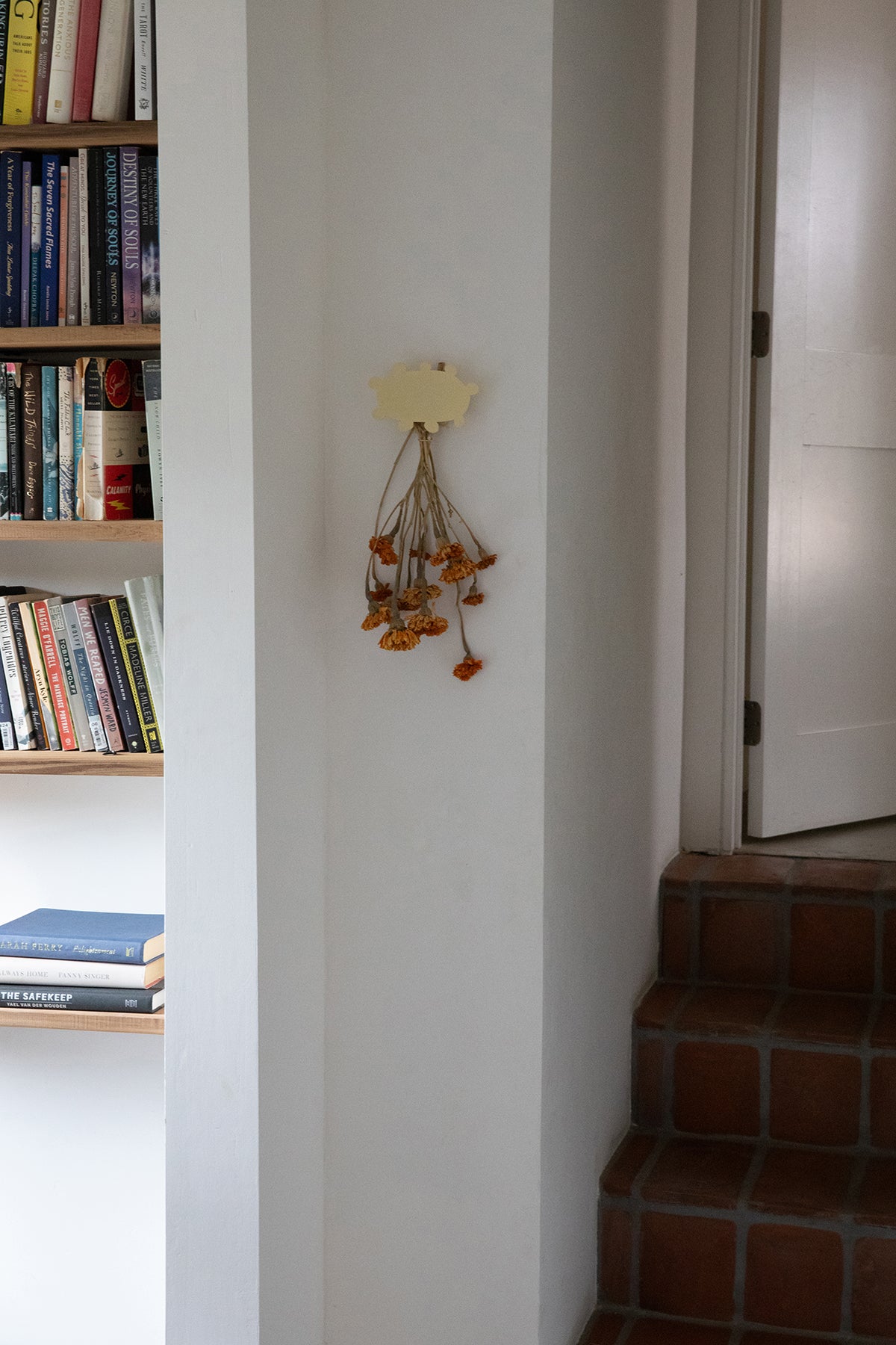 Upton Soli Wall Hook in butter. A sculptural decorative steel wall hook with a butter yellow color scheme with a unique abstract shape, designed to hang bags, coats, towels, hats, and more. Shown here holding a bouquet of dried flowers next to a bookshelf and stairs.