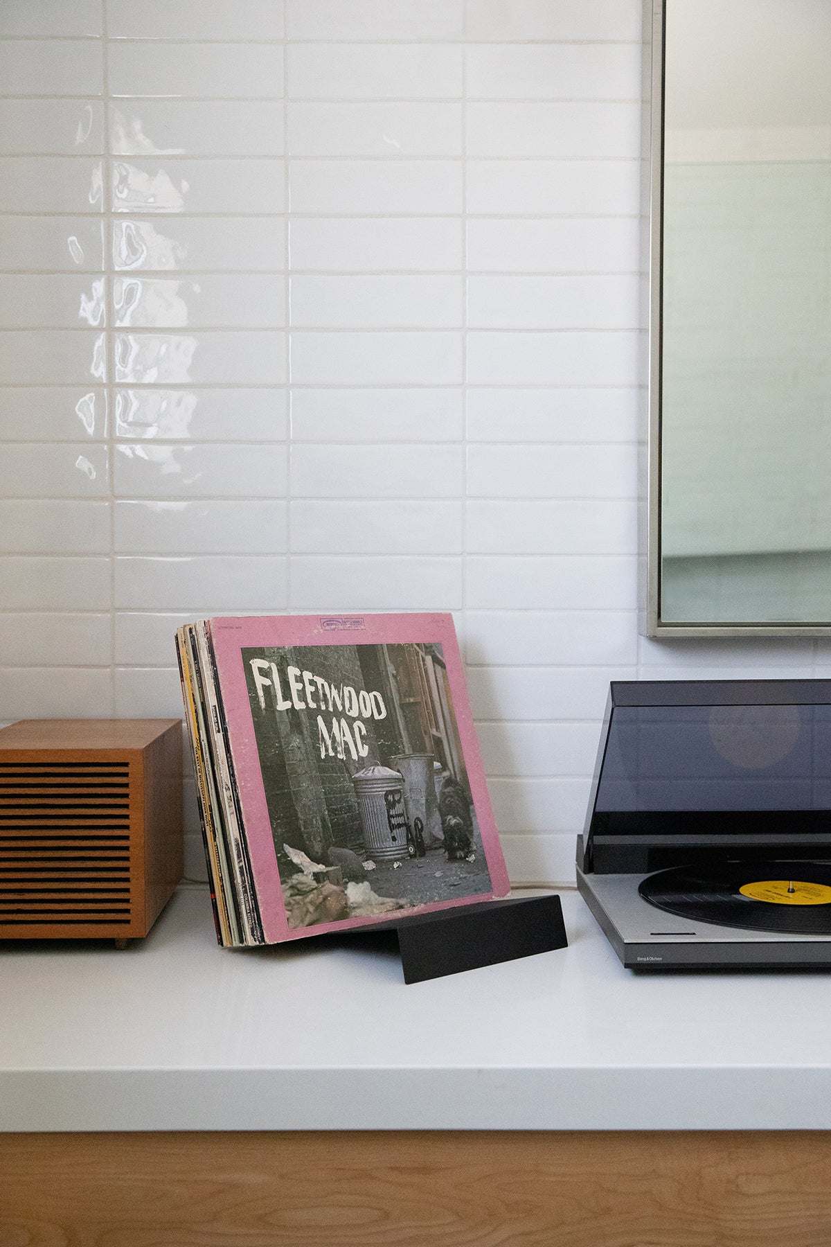 Upton LP Stand in black. Modern and minimalist tabletop steel vinyl record holder made for the storage, organization, and display 50 albums. Shown holding a Fleetwood Mac album with a record player on a white shelf with a tiled wall background in a modern interior.