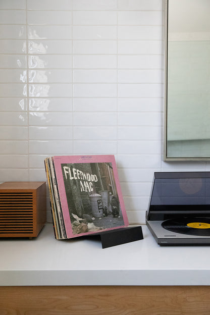 Upton LP Stand in black. Modern and minimalist tabletop steel vinyl record holder made for the storage, organization, and display 50 albums. Shown holding a Fleetwood Mac album with a record player on a white shelf with a tiled wall background in a modern interior.