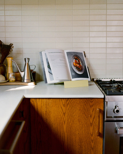 Upton LP Stand in pale yellow. Modern and minimalist tabletop steel vinyl record holder made for storage, organization, and display of 50 albums. Shown being used as a cookbook stand in a modern kitchen on a white shelf with wooden cabinets.
