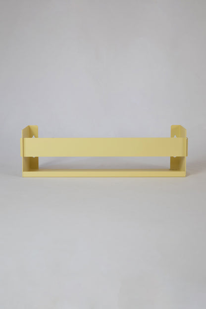 Upton Wall-Mounted Metal Shelf in butter yellow. With a bright, modern design and made from powder coated steel, this minimalist floating wall shelf is designed to be used as a bookshelf, magazine rack, cookbook holder, spice rack, knick knack shelf, or anything else you can think up. Shown on a white background.