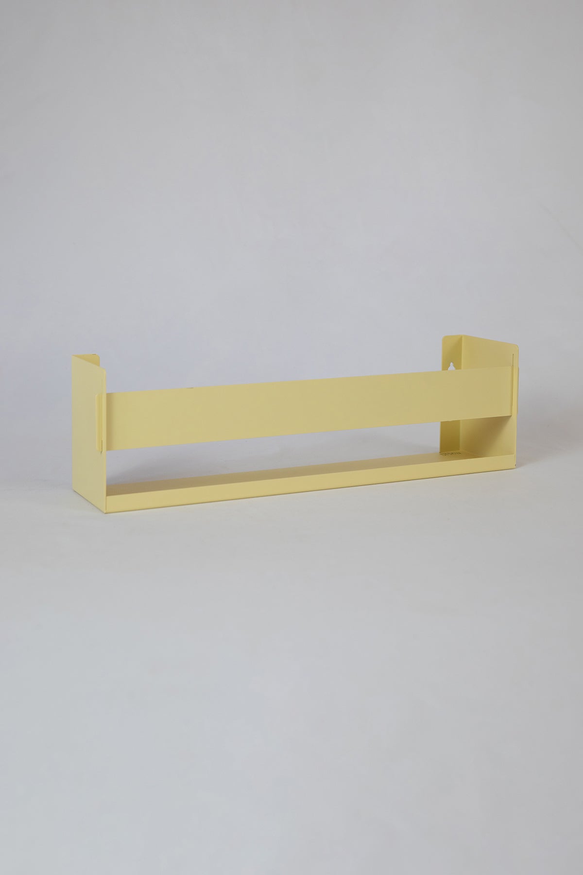 Upton Wall-Mounted Metal Shelf in butter yellow. With a bright, modern design and made from powder coated steel, this minimalist floating wall shelf is designed to be used as a bookshelf, magazine rack, cookbook holder, spice rack, knick knack shelf, or anything else you can think up. Shown on a white background.