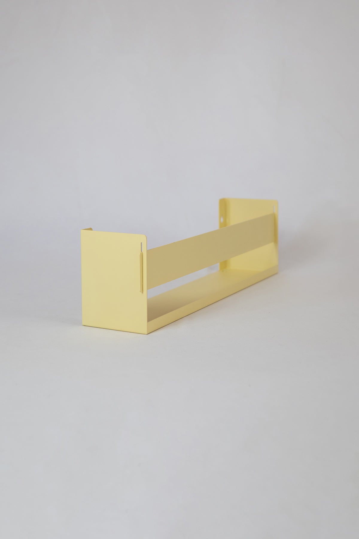 Upton Wall-Mounted Metal Shelf in butter yellow. With a bright, modern design and made from powder coated steel, this minimalist floating wall shelf is designed to be used as a bookshelf, magazine rack, cookbook holder, spice rack, knick knack shelf, or anything else you can think up. Shown on a white background.
