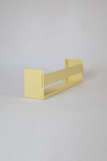 Upton Wall-Mounted Metal Shelf in butter yellow. With a bright, modern design and made from powder coated steel, this minimalist floating wall shelf is designed to be used as a bookshelf, magazine rack, cookbook holder, spice rack, knick knack shelf, or anything else you can think up. Shown on a white background.