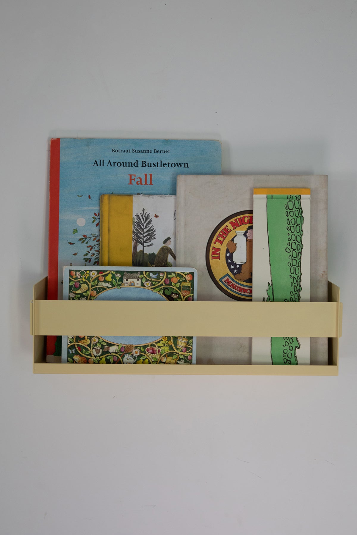 Upton Wall-Mounted Metal Shelf in butter yellow. With a bright, modern design and made from powder coated steel, this minimalist floating wall shelf is designed to be used as a bookshelf, magazine rack, cookbook holder, spice rack, knick knack shelf, or anything else you can think up. Shown holding several colorful books and printed items against a plain light gray wall.