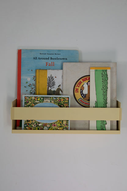 Upton Wall-Mounted Metal Shelf in butter yellow. With a bright, modern design and made from powder coated steel, this minimalist floating wall shelf is designed to be used as a bookshelf, magazine rack, cookbook holder, spice rack, knick knack shelf, or anything else you can think up. Shown holding several colorful books and printed items against a plain light gray wall.