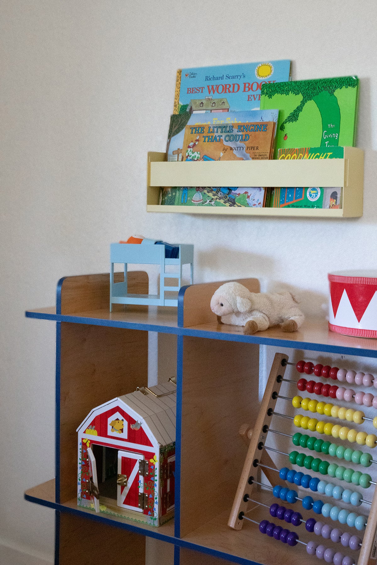 Upton Wall-Mounted Metal Shelf in butter yellow. With a bright, modern design and made from powder coated steel, this minimalist floating wall shelf is designed to be used as a bookshelf, magazine rack, cookbook holder, spice rack, knick knack shelf, or anything else you can think up. Shown above a wooden children's toy storage shelf with blue metal framing, styled with books on a top shelf, a small dollhouse, a stuffed bunny, a toy barn, and a colorful abacus.