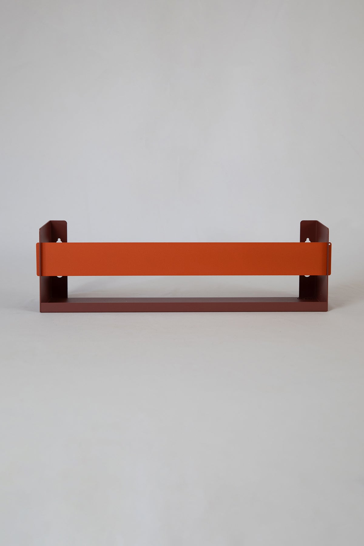Upton Wall-Mounted Metal Shelf in rose (red/pink) and burnt orange. With a bright, modern design and made from powder coated steel, this minimalist floating wall shelf is designed to be used as a bookshelf, magazine rack, cookbook holder, spice rack, knick knack shelf, or anything else you can think up. Shown holding several magazines against a muted green wall.