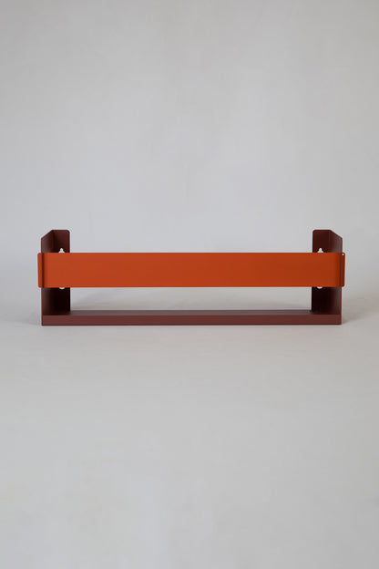 Upton Wall-Mounted Metal Shelf in rose (red/pink) and burnt orange. With a bright, modern design and made from powder coated steel, this minimalist floating wall shelf is designed to be used as a bookshelf, magazine rack, cookbook holder, spice rack, knick knack shelf, or anything else you can think up. Shown holding several magazines against a muted green wall.