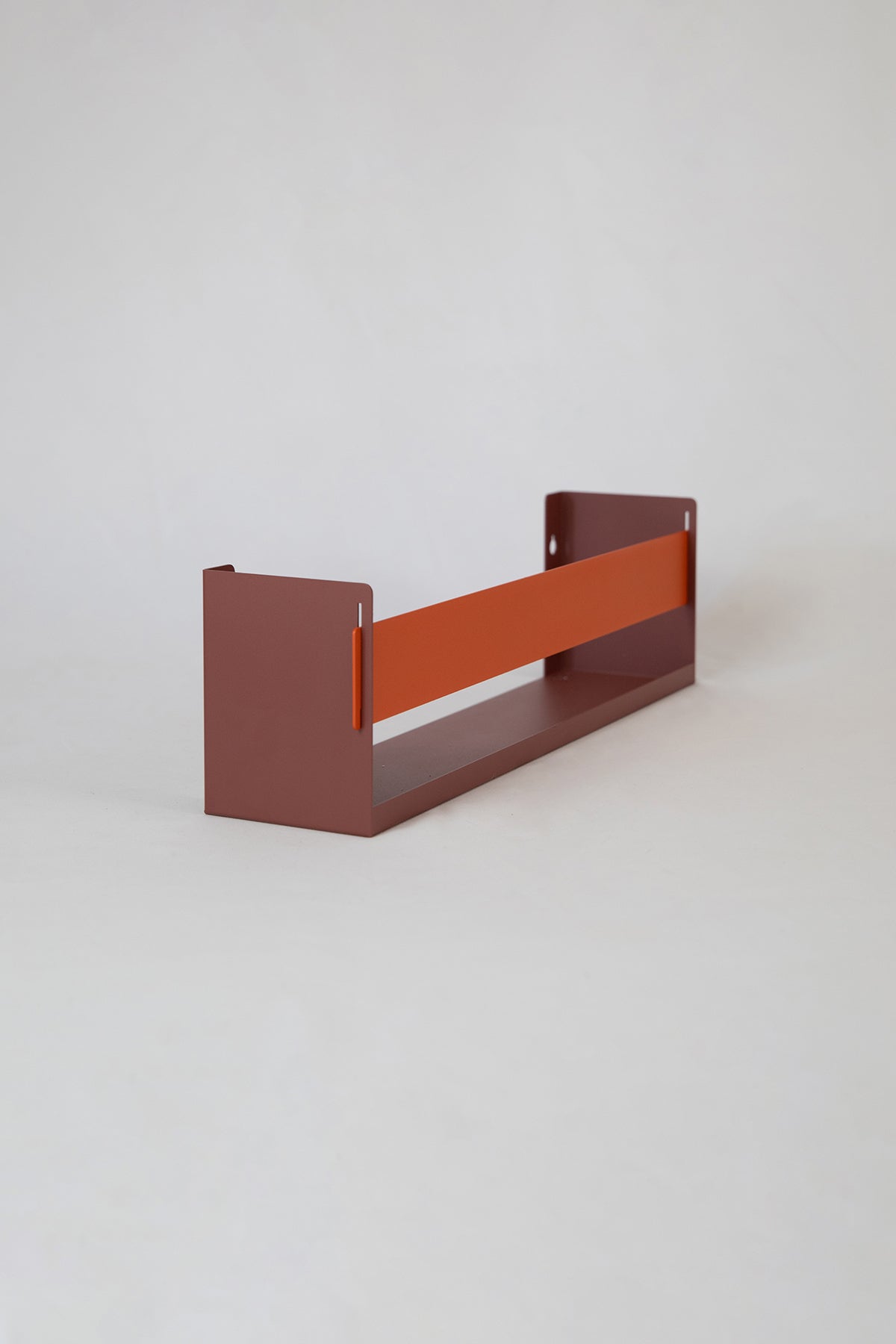 Upton Wall-Mounted Metal Shelf in rose (red/pink) and burnt orange. With a bright, modern design and made from powder coated steel, this minimalist floating wall shelf is designed to be used as a bookshelf, magazine rack, cookbook holder, spice rack, knick knack shelf, or anything else you can think up. Shown holding several magazines against a muted green wall.