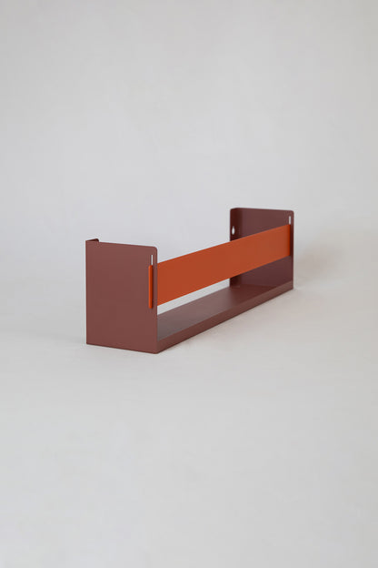 Upton Wall-Mounted Metal Shelf in rose (red/pink) and burnt orange. With a bright, modern design and made from powder coated steel, this minimalist floating wall shelf is designed to be used as a bookshelf, magazine rack, cookbook holder, spice rack, knick knack shelf, or anything else you can think up. Shown holding several magazines against a muted green wall.
