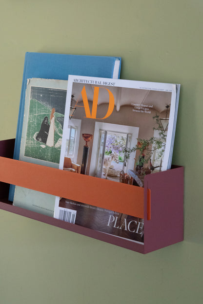 Upton Wall-Mounted Metal Shelf in rose (red/pink) and burnt orange. With a bright, modern design and made from powder coated steel, this minimalist floating wall shelf is designed to be used as a bookshelf, magazine rack, cookbook holder, spice rack, knick knack shelf, or anything else you can think up. Shown holding several magazines against a muted green wall.