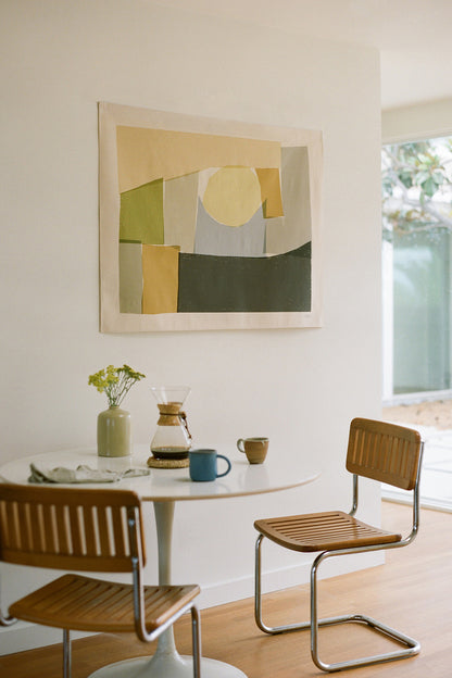 The Burren is an original art print on 100% cotton canvas from Michael Upton. Featuring an abstract block print composition in muted colors, the Burren is a large format modern wall hanging. Shown here in a modern sunny interior with mid century dining table and pour over coffee and ceramic vase with flowers.