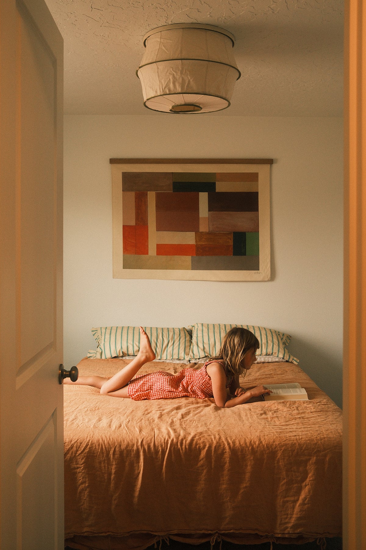 The Jardi is an original art print on 100% cotton canvas from Michael Upton. Featuring an architectural abstract design in warm earth tones, the Jardi is a large format modern wall hanging. Featured here in a cozy bedroom with a little girl reading under a warm light on a rust colored bedding.