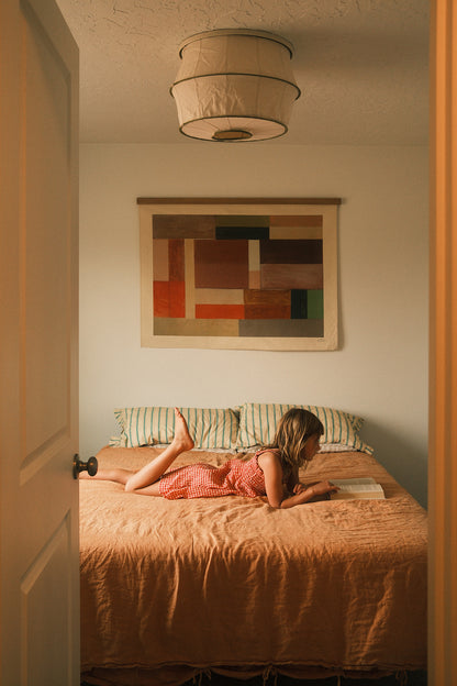 The Jardi is an original art print on 100% cotton canvas from Michael Upton. Featuring an architectural abstract design in warm earth tones, the Jardi is a large format modern wall hanging. Featured here in a cozy bedroom with a little girl reading under a warm light on a rust colored bedding.