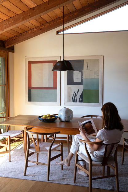 The Lark is an original art print on 100% cotton canvas from Michael Upton. Featuring an abstract collage design in rich green and blue hues, the Lark is a large format modern wall hanging. Shown here with a woman sitting at a wooden dining table in a modern room with handmade ceramics.