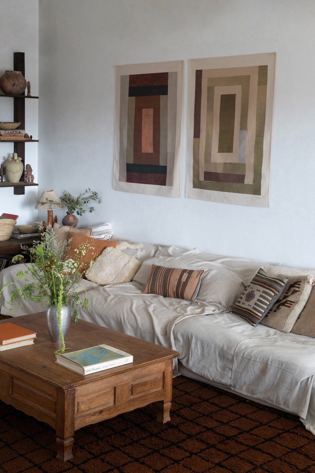 The Illum is an original art print on 100% cotton canvas from Michael Upton. Featuring an abstract quilt-like collage design in muted earth tones, the Illum is a large format modern wall hanging. Shown here in a living room with a white sofa and wooden coffee table.