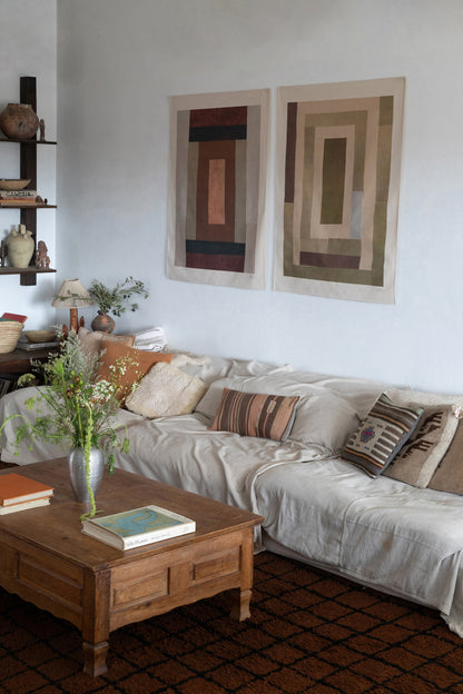 The Illum is an original art print on 100% cotton canvas from Michael Upton. Featuring an abstract quilt-like collage design in muted earth tones, the Illum is a large format modern wall hanging. Shown here in a living room with a white sofa and wooden coffee table.