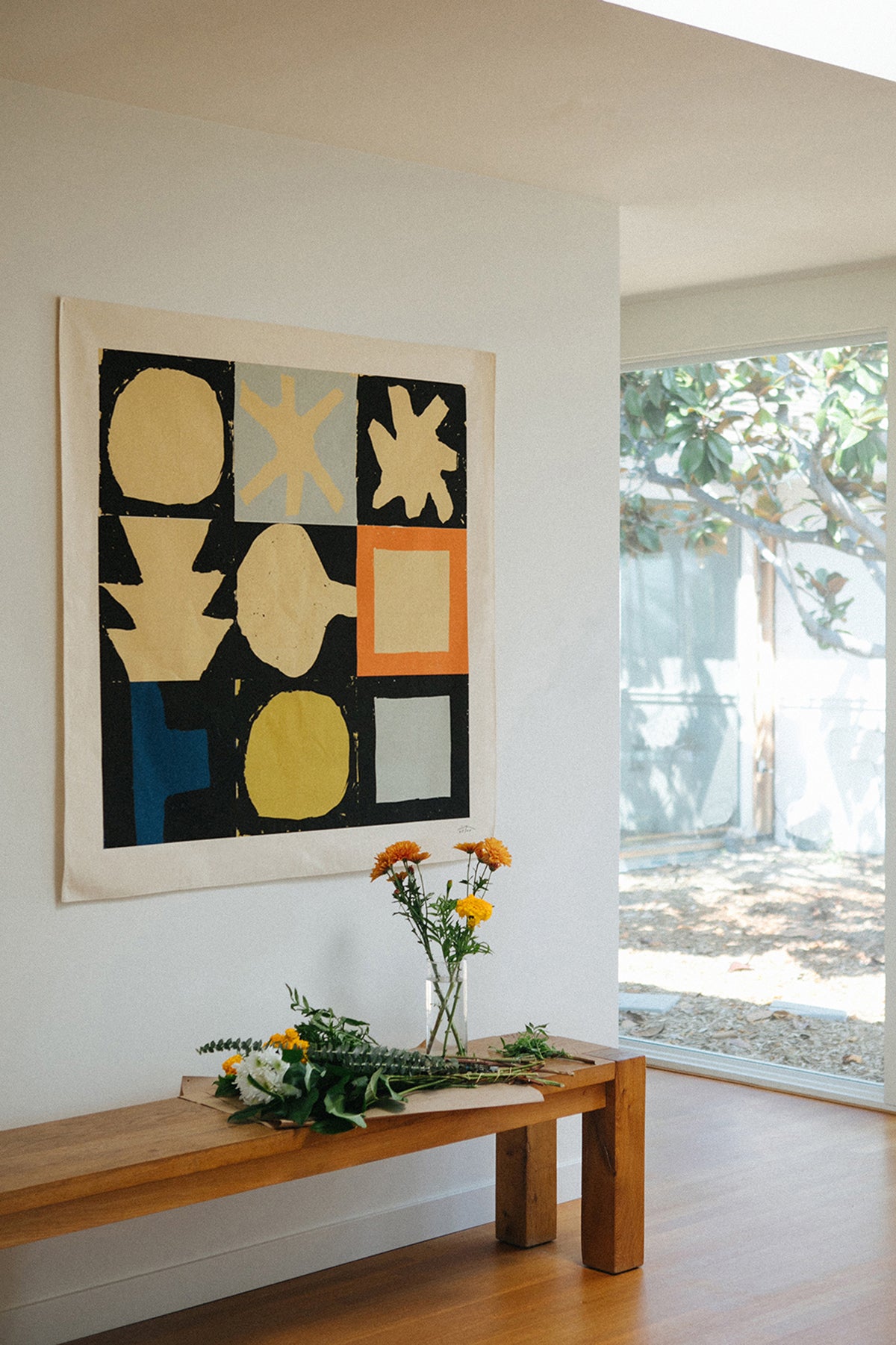 The Tenby is an original art print on 100% cotton canvas from Michael Upton. Featuring a geometric abstract mosaic design with bold colorful shapes, the Tenby is a large format modern wall hanging. Shown here in a modern interior with wooden bench and vase of flowers.