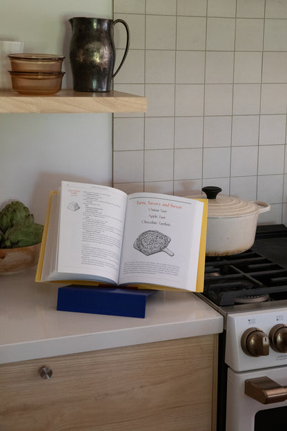 Upton LP Record Stand in cobalt blue. Modern and minimalist tabletop steel vinyl record holder made for the storage, organization, and display 50 albums. Shown here being used as a cookbook holder with an open copy of Alice Waters' "The Art of Simple Food" on a kitchen counter, with a stove, white tile backsplash, and open wooden shelf in the background.