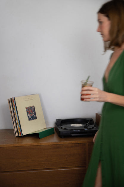 Upton LP Stand in pine green. Upton LP Stand in burnt orange. Modern and minimalist tabletop steel vinyl record holder made for storage, organization, and display of 50 albums. Shown holding a stack of records on a wooden table with record player while a woman in a green dress with cocktail in hand walks by.