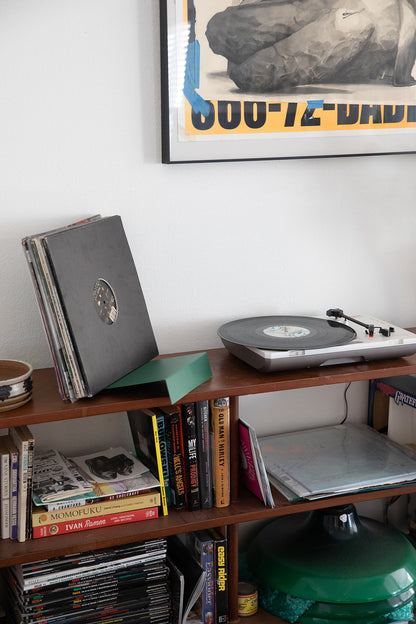 Upton LP Stand in pine green. Modern and minimalist tabletop steel vinyl record holder made for storage, organization, and display of 50 albums. Shown holding a stack of albums on a shelf with books and record player in an interior.