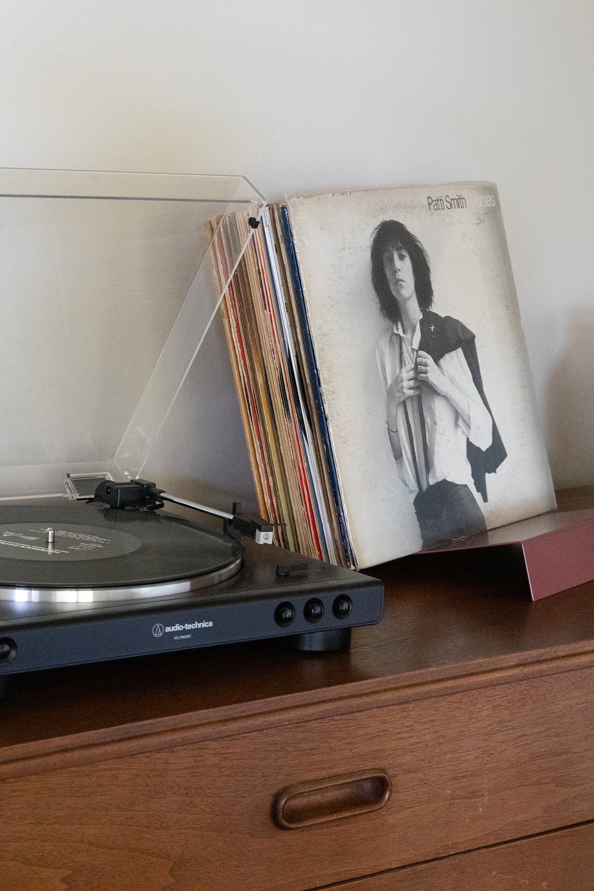 Upton LP Stand in rose (pink/red). Modern and minimalist tabletop steel vinyl record holder made for storage, organization, and display of 50 albums. Shown holding a Patti Smith album next to a record player on a wooden table.