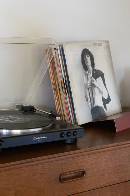 Upton LP Stand in rose (pink/red). Modern and minimalist tabletop steel vinyl record holder made for storage, organization, and display of 50 albums. Shown holding a Patti Smith album next to a record player on a wooden table.