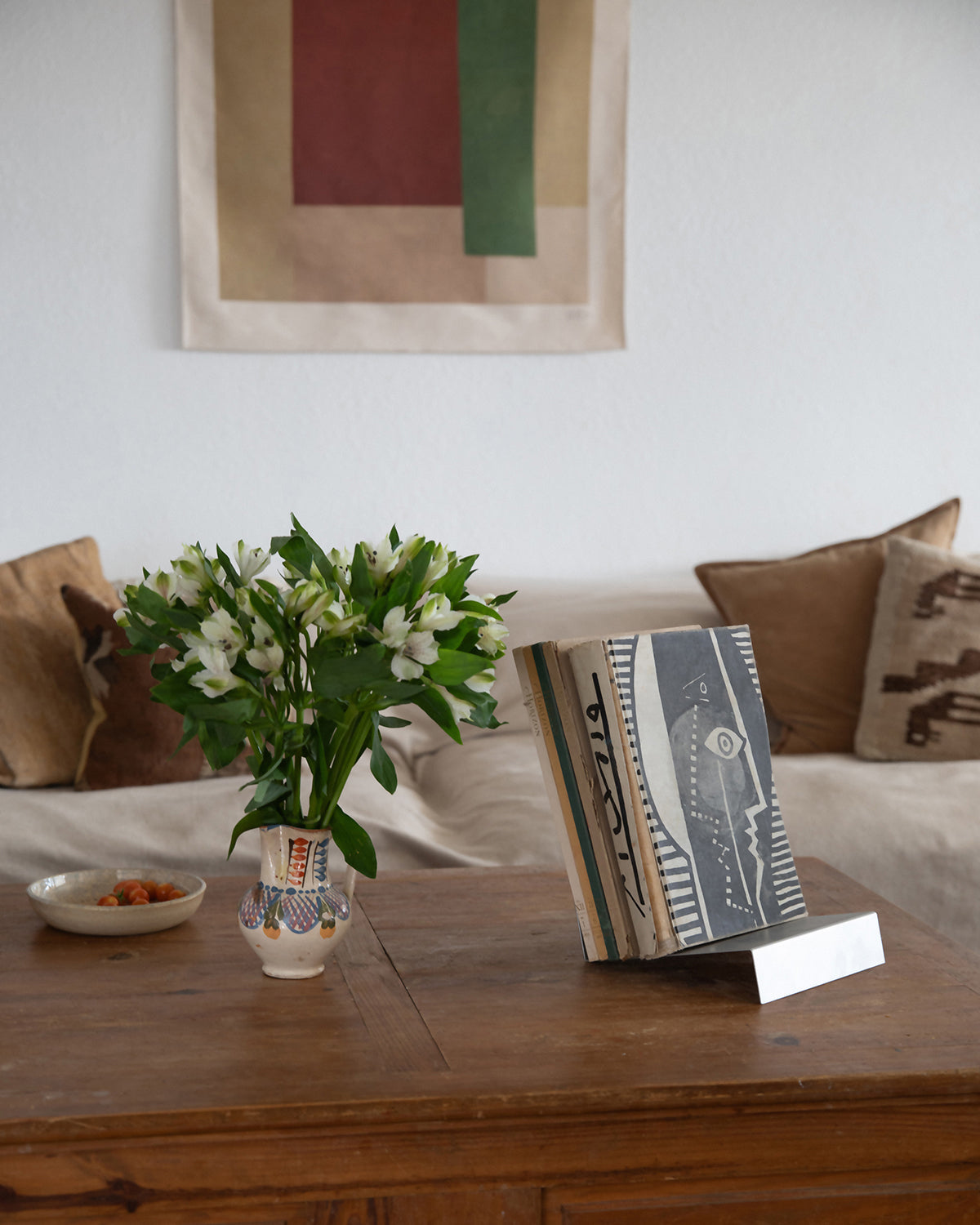 Upton LP Stand in matte silver. Modern and minimalist tabletop steel vinyl record holder made for storage, organization, and display of 50 albums. Shown being used as a tabletop book stand in a rustic interior holding a stack of art books on a wooden table with flowers.