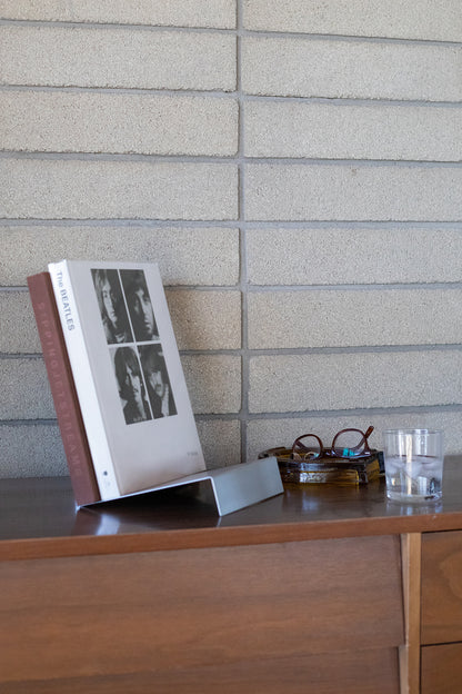 Upton LP Stand in matte silver. Modern and minimalist tabletop steel vinyl record holder made for storage, organization, and display of 50 albums. Shown being used as a tabletop book stand holding a stack of art books on a wooden shelf with water glass and glasses in a modern interior.