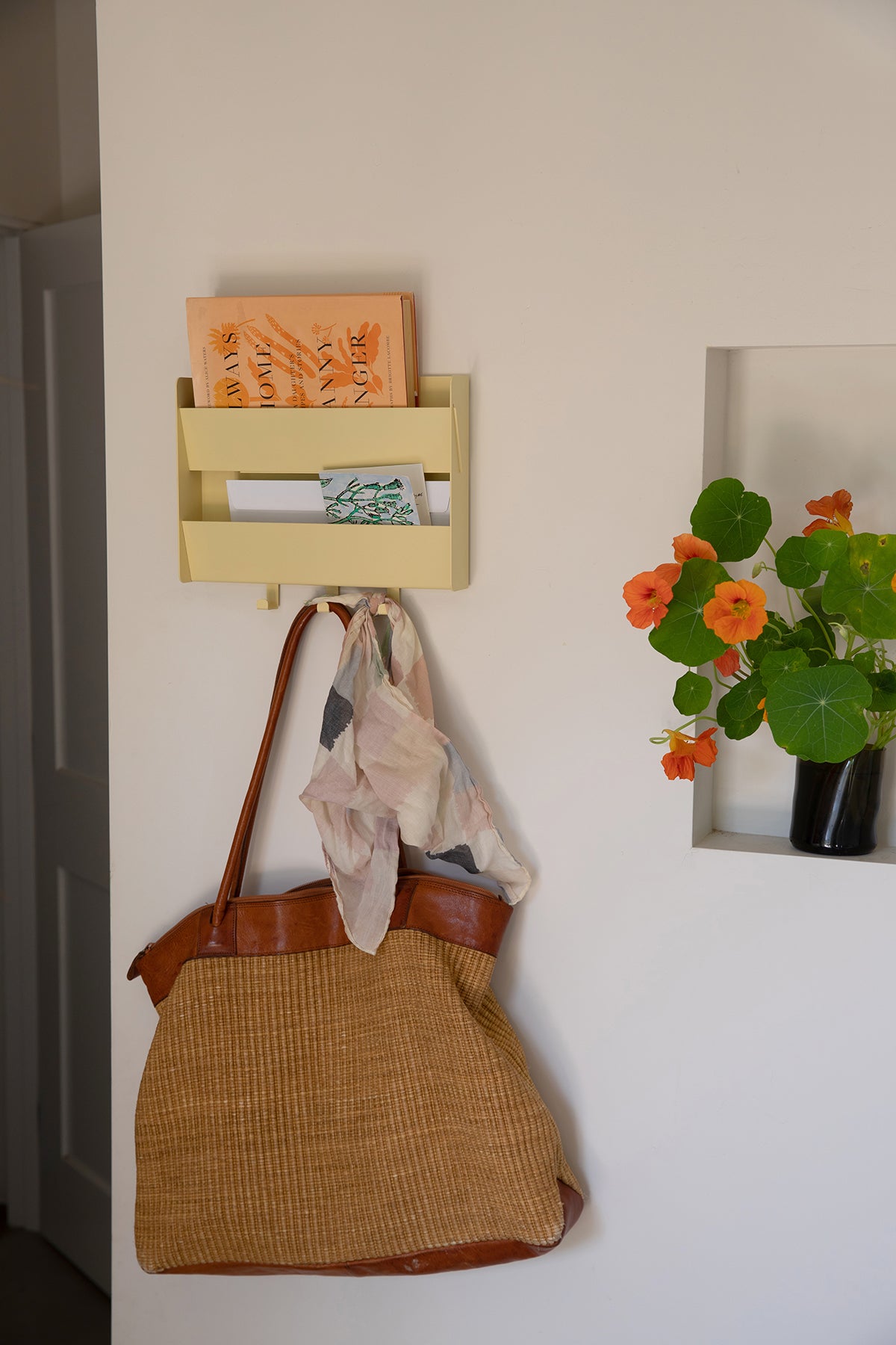 The Upton Mail Organizer in butter. Modern and design-forward wall-mounted mail organizer made of steel with a light yellow powder coat finish. Featuring two slots for mail and three sturdy hooks. Shown here on a white wall holding a Fanny Singer book, pink headscarf, and tote bag next to a vase of nastirtium flowers.