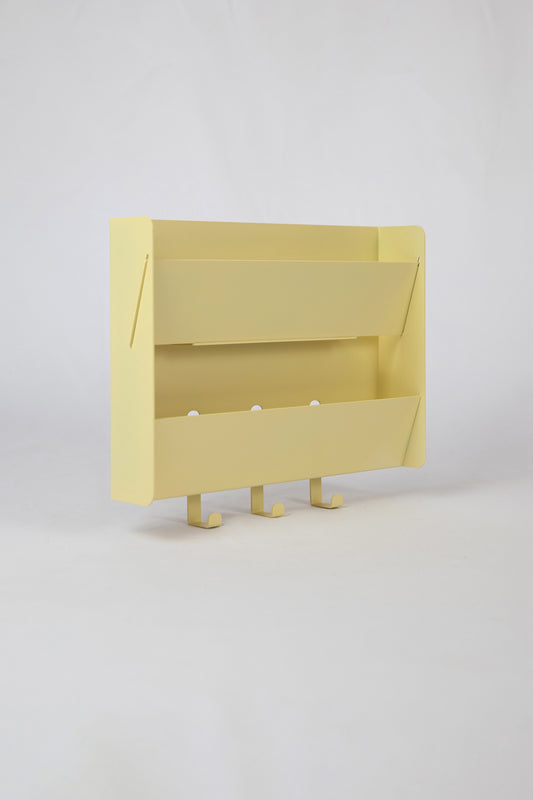The Upton Mail Organizer in butter. Modern, minimalist, and design-forward wall-mounted mail organizer made of steel with a light yellow powder coat finish. Featuring two slots for mail and three sturdy hooks to hang keys, coats, bags, etc. Shown here on a white background.