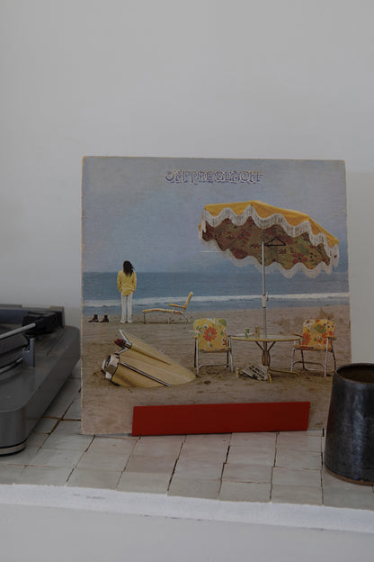 Upton Now Playing Record Stand in brick red (red orange). Modern and minimalist steel display stand made to hold the vinyl record sleeve for the vinyl album currently playing on the turntable. Shown holding an "On the Beach" album featuring a cover with a beach scene with a yellow umbrella, lounge chair, table, and a person standing by the ocean, displayed upright on a white tile shelf against a plain wall.