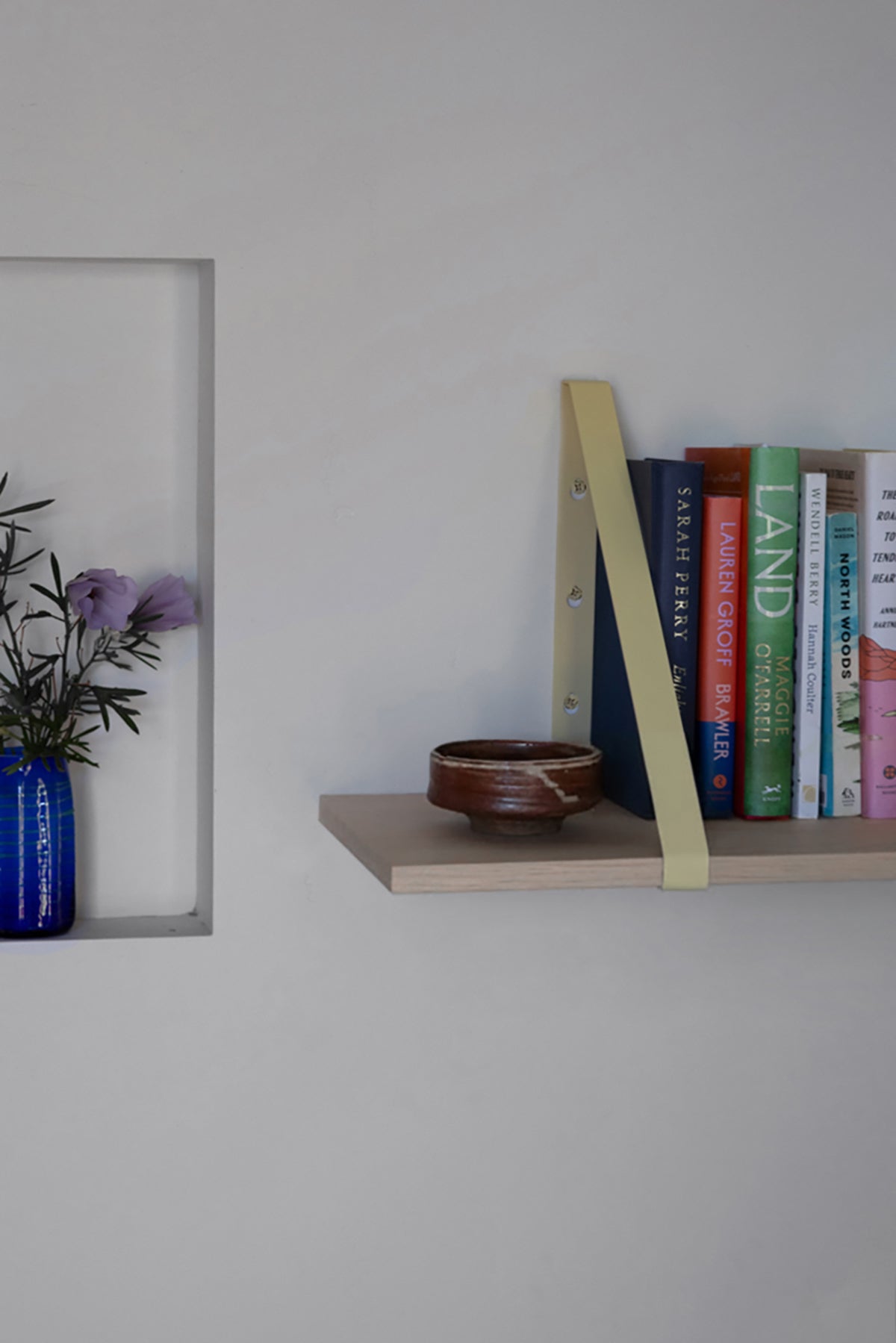 Upton Shelf Bracket Set in butter yellow. With a modern, minimalist design and made from powder coated steel, these slim triangle brackets are a unique home storage solution that adds character and color to your home. Shown in a minimalist interior scene being used to make a light wood floating shelf mounted on a white wall. The shelf holds a small brown ceramic bowl and a row of colorful books.