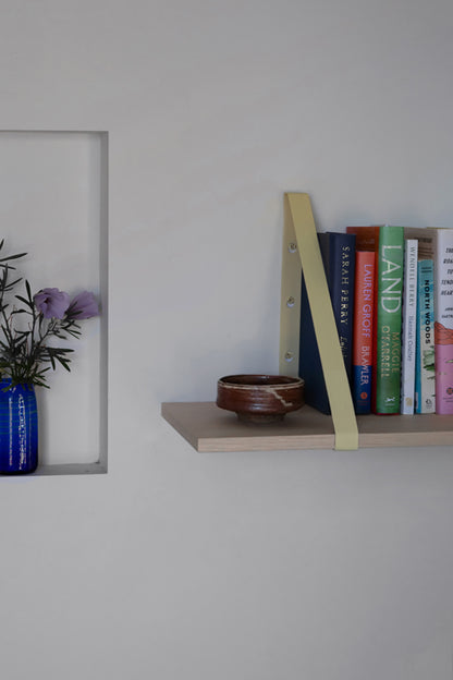 Upton Shelf Bracket Set in butter yellow. With a modern, minimalist design and made from powder coated steel, these slim triangle brackets are a unique home storage solution that adds character and color to your home. Shown in a minimalist interior scene being used to make a light wood floating shelf mounted on a white wall. The shelf holds a small brown ceramic bowl and a row of colorful books.