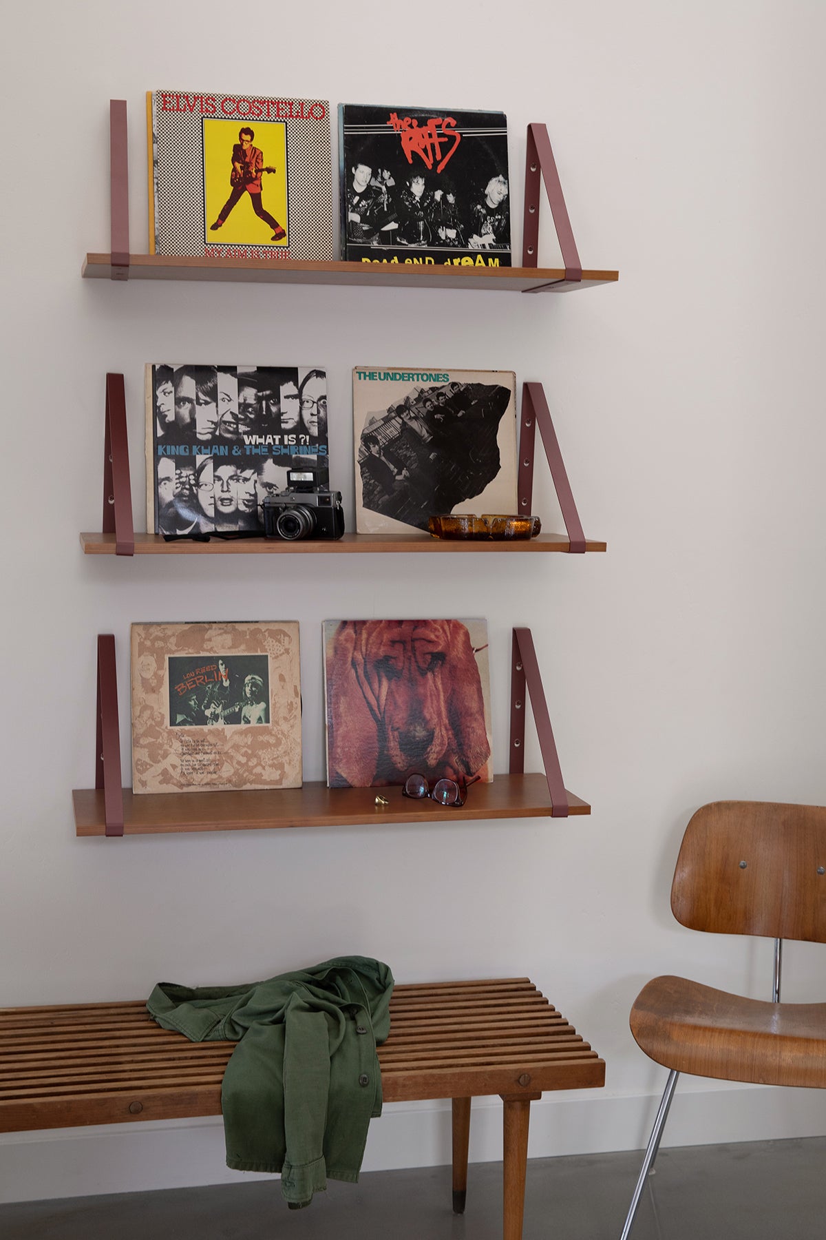 Upton Shelf Bracket Set in rose (red/pink). With a modern, minimalist design and made from powder coated steel, these slim triangle brackets are a unique home storage solution that adds character and color to your home. Shown being used to make three wooden floating shelves mounted on a white wall. The shelves display various vinyl records, a small bowl, and a framed print.