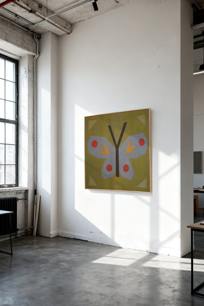 Butterfly Stamp by Sunroom Studios x Upton, a custom framed art print on 100% cotton canvas. Large cut paper collage of a butterfly on an olive green background, with light blue, red, and yellow accents. Originally created as a risograph print. Shown on a white wall in a sunlit industrial loft with concrete floors and exposed ceiling beams.