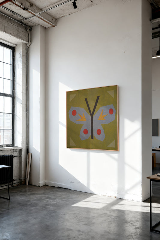 Butterfly Stamp by Sunroom Studios x Upton, a custom framed art print on 100% cotton canvas. Large cut paper collage of a butterfly on an olive green background, with light blue, red, and yellow accents. Originally created as a risograph print. Shown on a white wall in a sunlit industrial loft with concrete floors and exposed ceiling beams.