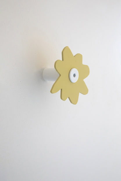 Upton Fleur Wall Hook in pale yellow. A sculptural decorative steel wall hook with a pale yellow color scheme with a unique abstract flower shape, designed to hang bags, coats, towels, hats, and more. Shown here on a white background.