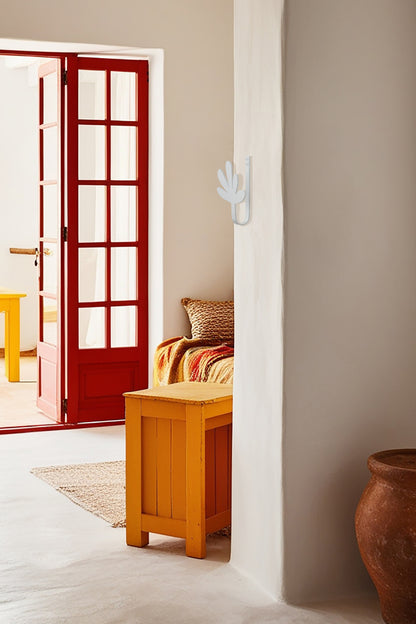 Upton Flora Wall Hook in white. A sculptural decorative steel wall hook with a white color scheme with a unique abstract shape, designed to hang bags, coats, towels, hats, and more. Shown here in a rustic interior with red door, orange furniture and decorative vase.