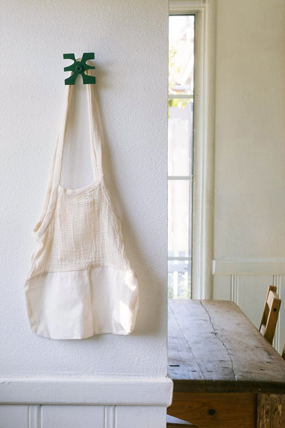 Upton Klipp Wall Hook in pine. A sculptural decorative steel wall hook with a pine green color scheme with a unique abstract shape, designed to hang bags, coats, towels, hats, and more. Shown here holding a white mesh famers market tote bag in a rustic interior with wooden dining table and white walls.