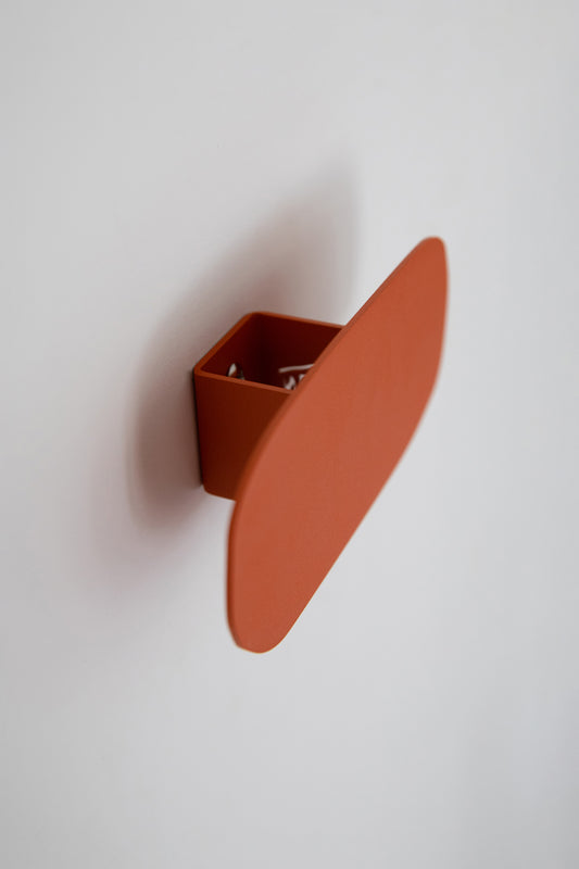 Upton Pill Wall Hook in burnt orange. A sculptural decorative steel wall hook with a burnt orange color scheme with a unique abstract shape, designed to hang bags, coats, towels, hats, and more. Shown here on a white background.
